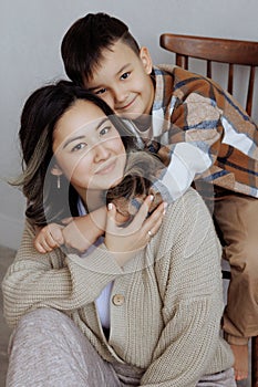 Mother and child in cozy casual clothes pose on a wooden chair. Studio shot.