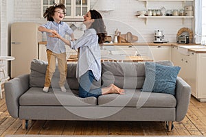Mother and child laughing having fun, holding hands and jumping on sofa at home. Happy family.