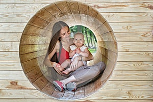 Mother and child are sitting inside wooden tunnel on the Playground. Happy mom and baby