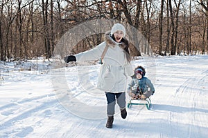 Mother and child sledge in the snowy forest from the mountain. Family vacation on the street in winter, mom and son laugh and have