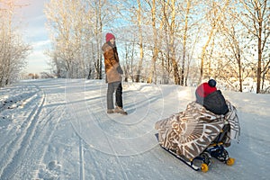 Mother with a child walks on a frosty winter morning on a Sunny day. Baby wrapped in a blanket rides on a sled