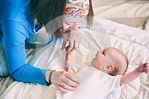 Mother and child on a white bed. Mom and baby boy in diaper playing in sunny bedroom. Mom makes gymnastics for her newborn baby.