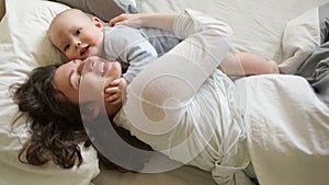 Mother and child on a white bed. Mom and baby boy in diaper playing in sunny bedroom. Parent and little kid relaxing at