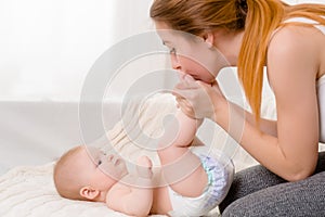 Mother and child on a white bed. Mom and baby girl in diaper playing in sunny bedroom.