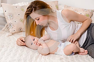 Mother and child on a white bed. Mom and baby girl in diaper playing in sunny bedroom.