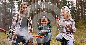 mother with children frying and eating sausages by bonfire while camping in forest. family time, nature adventure