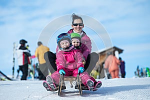 Mother and children having fun on sledge with panoramatic view of Alps mountains. Active mom and toddler kid with safety helmet.