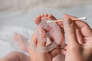 Mother cleaning baby`s foot with cotton bud on blurred background, closeup