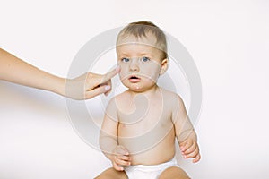 Mother creaming cute infant girl face. Little baby girl with cream on her face sitting on the white background