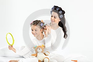 Mother in curlers curling hair to happy little daughter looking at hand mirror