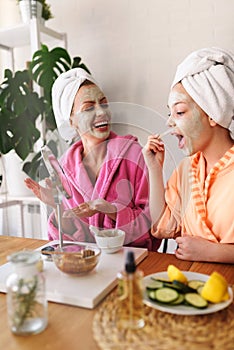 Mother and daughter in bathrobes and towels on head using natural cosmetics and having fun