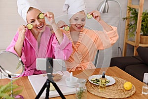 Mother and daughter in bathrobes and towels on head using natural cosmetics and having fun live online