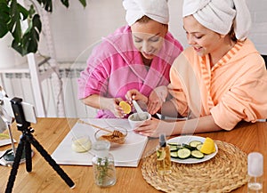 Mother and daughter in bathrobes and towels on head using natural cosmetics and having fun live online