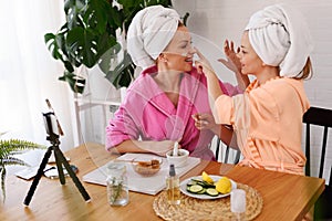 Mother and daughter in bathrobes and towels on head using natural cosmetics and having fun live online