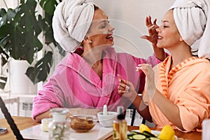 Mother and daughter in bathrobes and towels on head using natural cosmetics and having fun live online