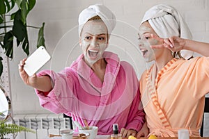 Mother and daughter in bathrobes and towels on head using natural cosmetics and having fun together at home