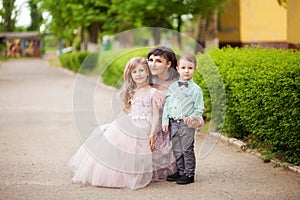 Mother with daughter in beige lush dresses and son in a green shirt are in the summer green park, a woman with two children