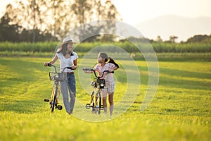 Mother and daughter with bicycling at the garden meadow in sunset near white fence.