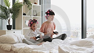 Mother and daughter in curlers sitting on bed at home and playing video games.