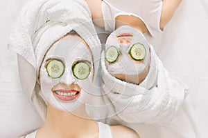 Mother and daughter doing funny spa procedures after bath. They are in white bath towels with white facial mud mask on