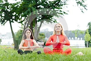 Mother and daughter doing yoga exercises on grass in the park at the day time. People having fun outdoors. Concept of friendly