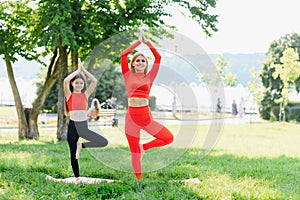 Mother and daughter doing yoga exercises on grass in the park at the day time. People having fun outdoors. Concept of friendly