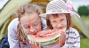 Mother and daughter eating watermelon near a tent in meadow or park. Happy family on picnic at camping