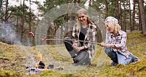 mother and daughter frying sausages over a campfire in the forest. family time