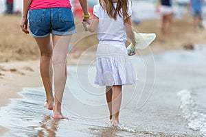 Mother and daughter girl walking together on sand beach in sea water in summer with bare feet in warm ocean waves