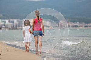 Mother and daughter girl walking together on sand beach in sea water in summer with bare feet in warm ocean waves