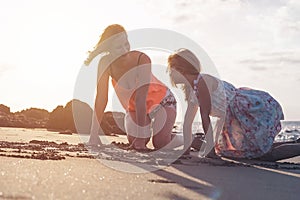 Mother and daughter having fun drawing with sand at sunset on tropical beach - Happy mom playing with her kid in vacation