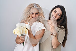 Mother and daughter holding bouquet of white flowers cheerful with a smile on face pointing with hand and finger up to the side