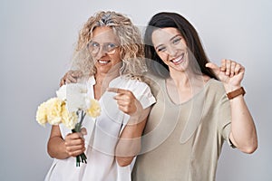 Mother and daughter holding bouquet of white flowers pointing to the back behind with hand and thumbs up, smiling confident