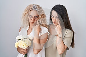Mother and daughter holding bouquet of white flowers pointing to the eye watching you gesture, suspicious expression