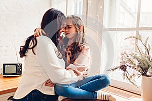 Mother and daughter hugging on kitchen window sill by decorated Christmas tree. Family spending time together at home