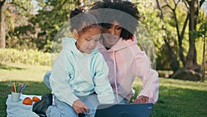 Mother daughter looking tablet on green meadow close up. African family picnic