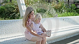 Mother and daughter moment, mum sitting on a park bench, putting shoes on her little girl outdoors amid lush green nature