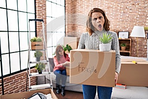 Mother and daughter moving to a new home holding cardboard box in shock face, looking skeptical and sarcastic, surprised with open