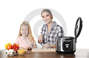 Mother and daughter preparing food with multi cooker at table against white background