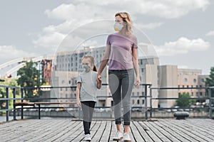 mother and daughter in protective masks holding hands on pier and looking away air