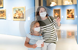 Mother and daughter in protective masks looking at paintings in halls of museum