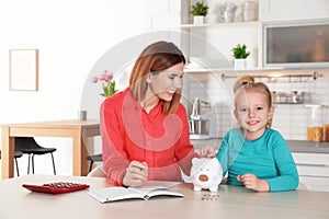 Mother and daughter putting coin into piggy bank at table indoors