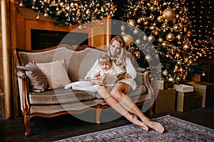 Mother and daughter reading a book at fireplace on Christmas eve. Decorated living room with tree, fire place and gifts. Winter