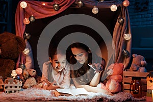 Mother and daughter are reading book with flashlight in pillow house late at night at home.
