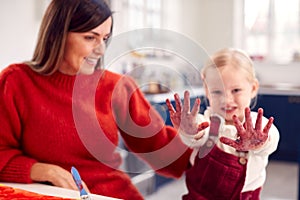 Mother With Daughter Showing Messy Hands At Home Doing Craft And Painting Picture In Kitchen