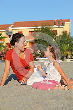 Mother with daughter sit on sand and laugh