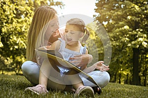 Mother and daughter sitting on green grass and reading book together. Little girl sitting on mother lap.