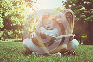 Mother and daughter sitting on green grass and reading book together. Little girl sitting on mother lap.