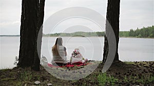 Mother and daughter are sitting on the shore of the lake between the pines and watching the water.