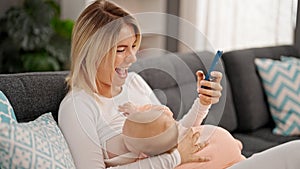 Mother and daughter sitting on sofa breastfeeding baby using smartphone at home